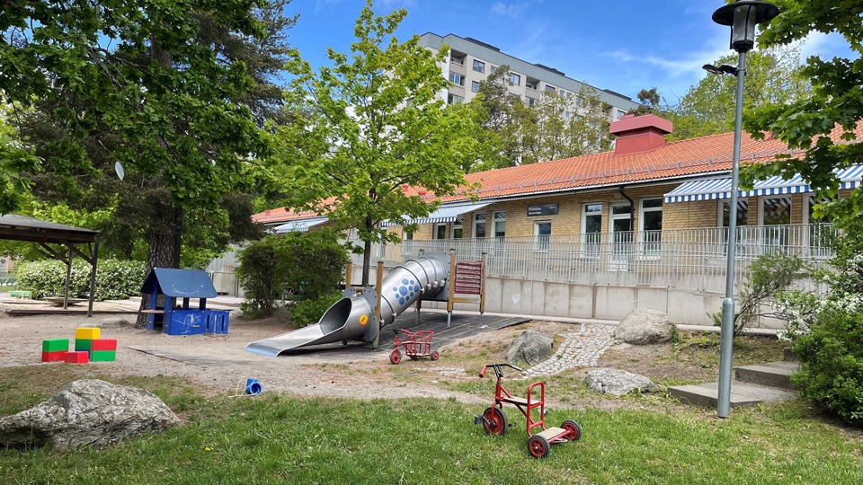Gul tegelbyggnad med grönskande gård där det finns leksaker, foto. 