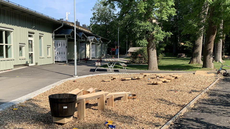 Förskola i bakgrunden med gård framför sig med lekhus, sandlåda, cyklar, bord att sitta vid och grönska.