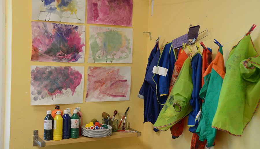 Målarfärg och målarkläder samt barnens målningar på väggen inne på förskolan.
