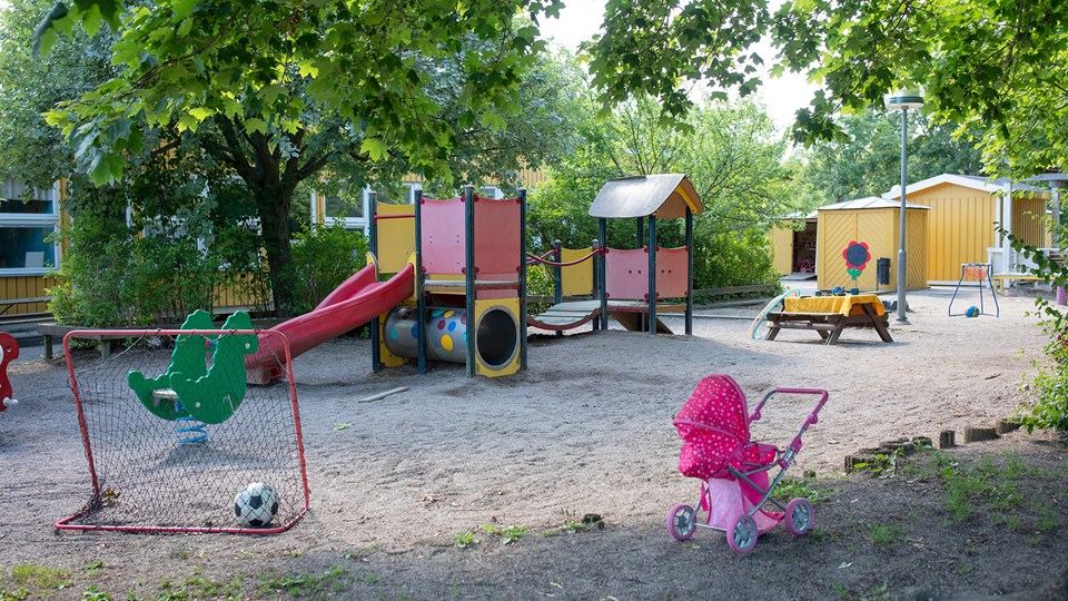 Förskolegård med buskar, träd och asfalt samt stor grusplan med klätterborg, fotbollsmål, pysselbord, dockvagn och andra leksaker.