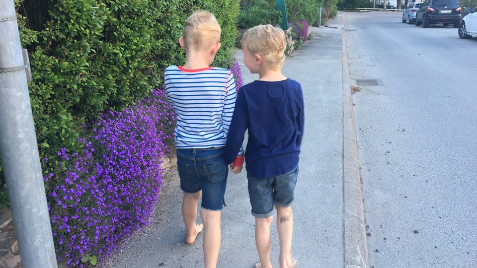 Två sommarklädda barn går och pratar på trottoaren. Blå blommor och gröna buskar.