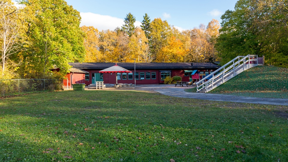 Byggnad med skog i bakgrunden samt gräsmatta, kulle, sandlåda och lekobjekt framför sig.