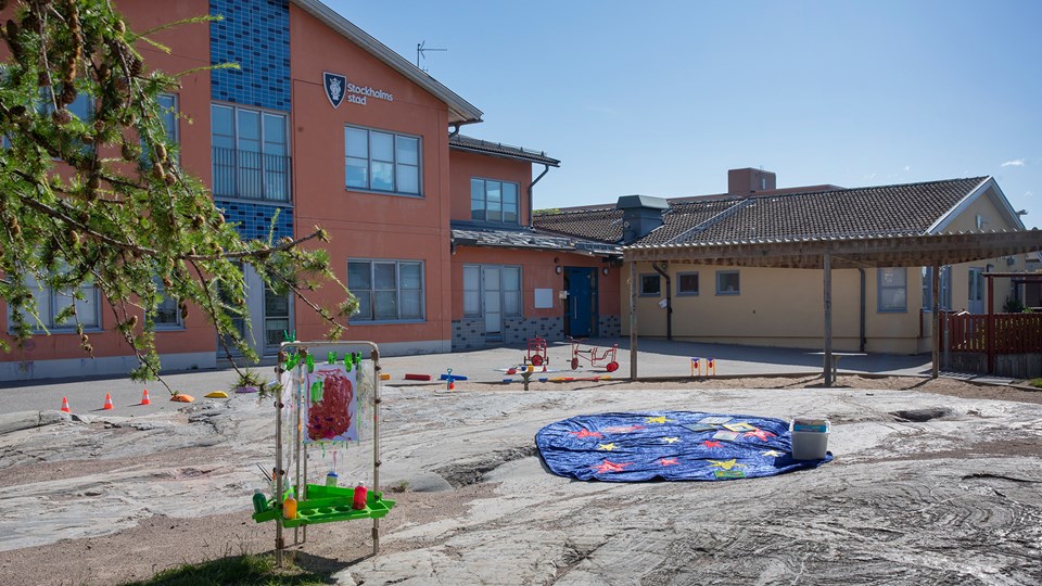 Förskolegård med träd, klippor, sandlåda och asfalt med trehjulingar och andra leksaker.