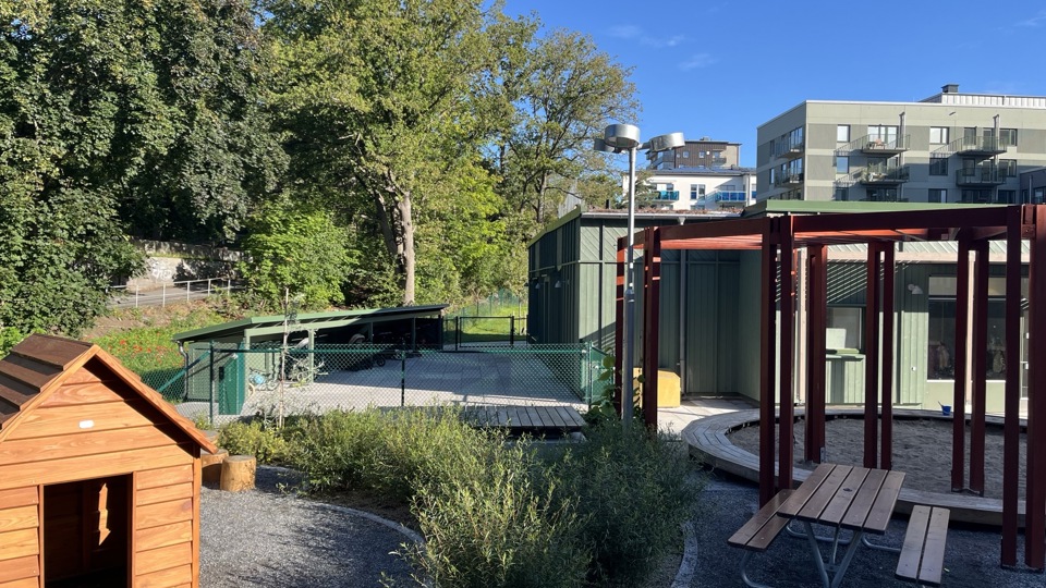 Lekyta med sittplats och minihus framför en grön förskolebyggnad. Grå flerfamiljshus syns i bakgrunden.