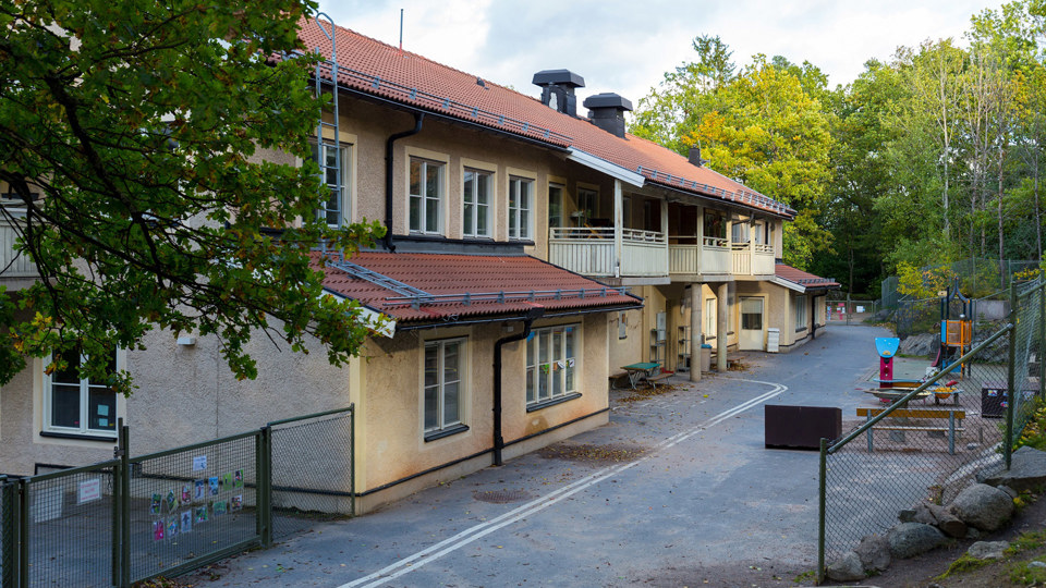 Förskolebyggnad med asfalterad gårdsplan och flera olika lekredskap. Träd och sluttning med stenar.