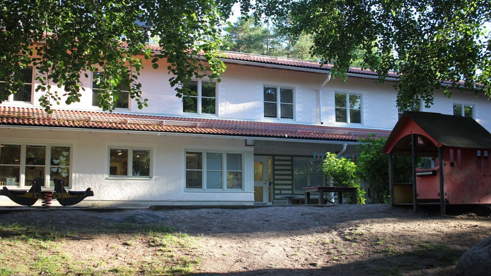Förskolan Fyrbåken, ett tvåplanshus i en putsad naturvit yta med röda tegelpannor. Intill står ett rött lekhus, en sittyta och en gunga i form av två sälar.