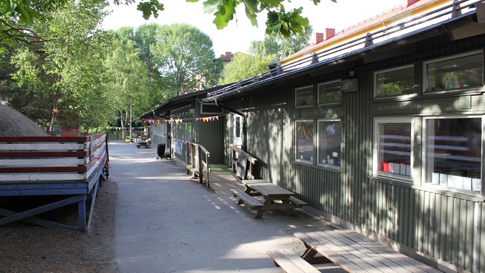 På högersida ser man förskolans gröna fasad med klassiska parkbänkar utanför. På vänstersida skymtar man förskolans gård.