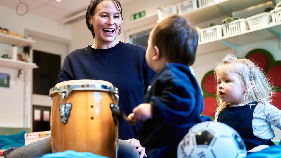 En vuxen person sitter och spelar på en kongatromma med två barn, ett av barnen håller i en fotboll. De ser glada ut i en lekmiljö.
