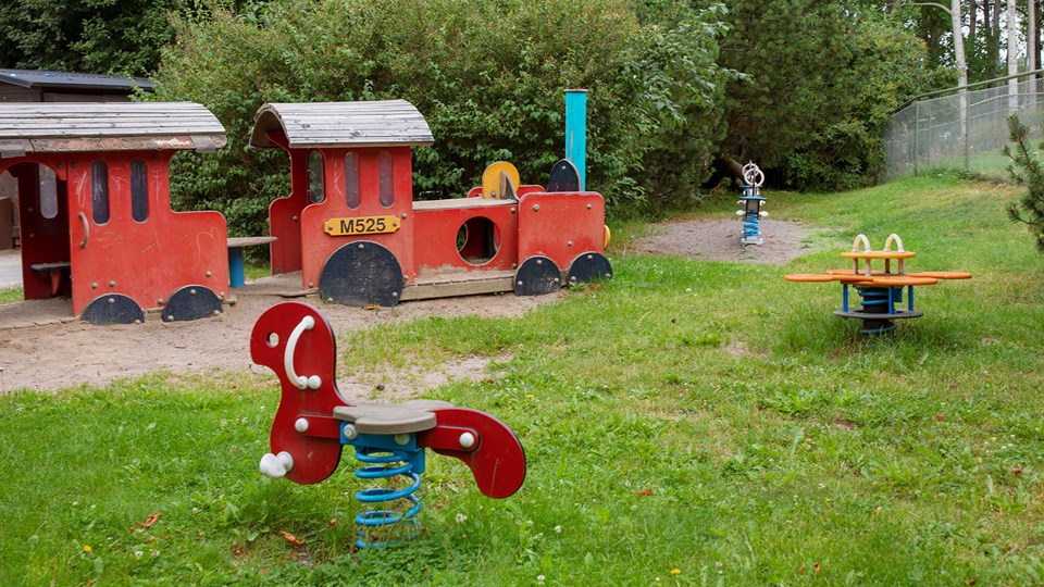 Del av förskolegård med buskar, träd och gräsmatta samt ett stort tåg och leksaker som man kan gunga på.