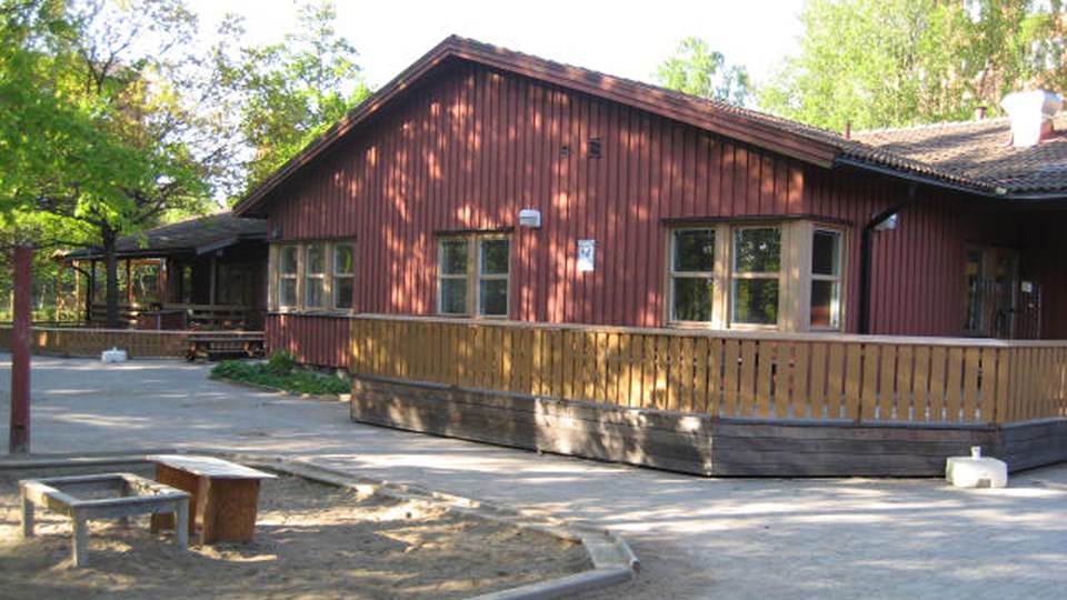 Förskolan Rödmyrans röda träfasade syns bakom ett gult staket. Man ser också en sandlåda i vänster kant.