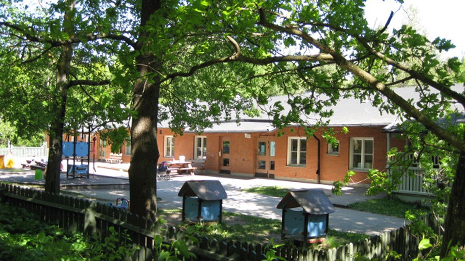 Förskolans rödbruna fasad skymtas mellan lummiga ekar. Delar av förskolegården syns i förgrunden med små lekhus och klätterställning.
