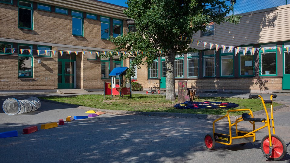 Förskolegård med träd, gräsmatta och asfalt med trehjulingar och andra leksaker.