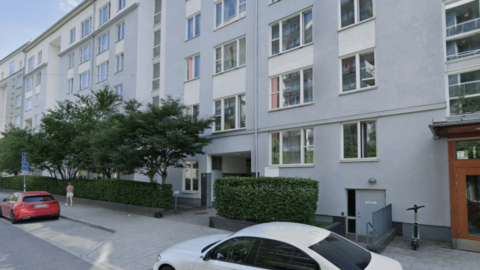 Utsidan av en förskola med ljus fasad, träd och parkerade bilar. En person går på trottoaren utanför.