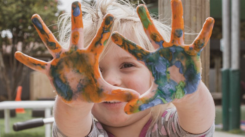 Ett barn sträcker leende fram sina händer som är fulla av målarfärg.