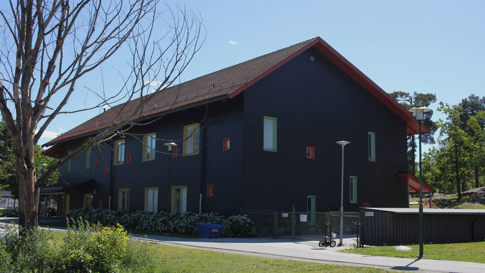 Förskolan Långe Erik, ett svart tvåplanshus med rött tak syns längs en gångväg. Bakom huset skymtas gården med berg och träd.