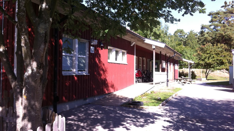 Röd förskolebyggnad med lummiga träd runt omkring och en grind i förgrunden.