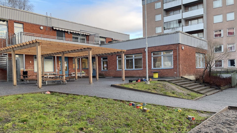 Förskolebyggnad med stor gård framför sig med gräsyta, sandlåda, asfalt och sittgrupper.