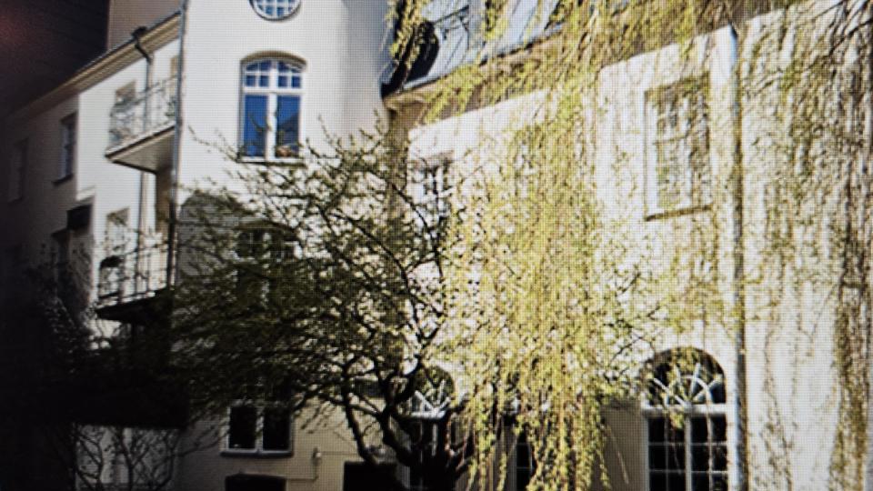 Ett stort vitt sekelskifteshus med runda och fyrkantiga former. Grönskande lövträd.