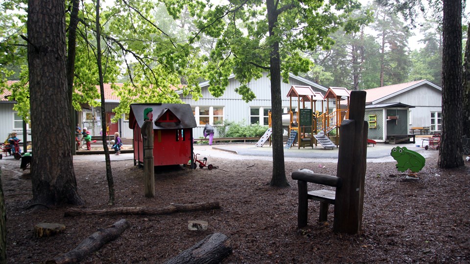 Förskolegården med lekredskap, träd och stockar, förskolans hus i bakgrunden.