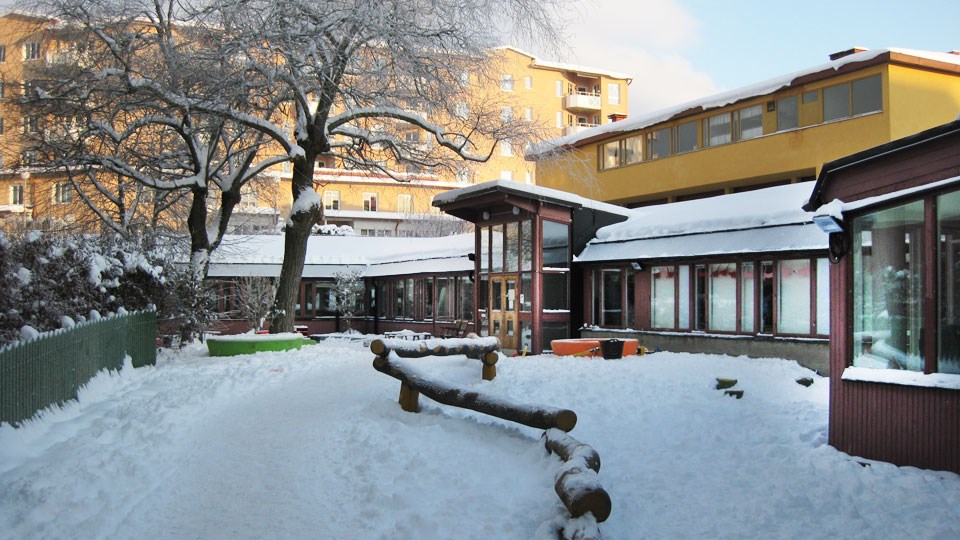 Bild på förskolan torget i vinterskrud. Förskolans gård i förgrunden. Klätterstockar sticker upp ur snön.
