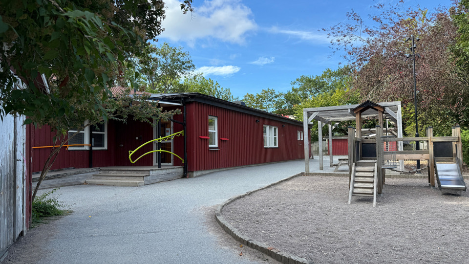 Röd förskolebyggnad med en asfalterad gång och en lekplats med klätterställning och rutschkana.