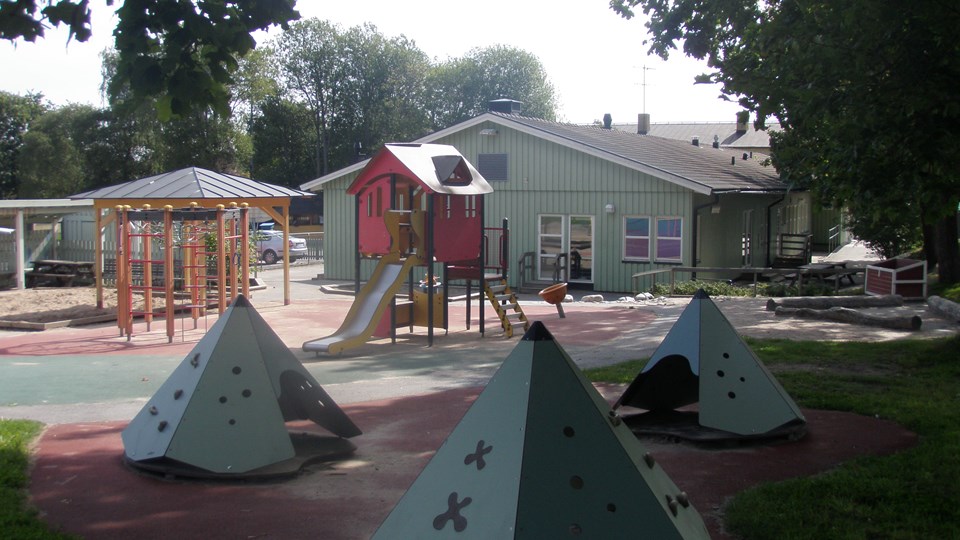 Grön förskolebyggnad i bakgrunden. I förgrunden ser man flera lekredskap i olika färger.