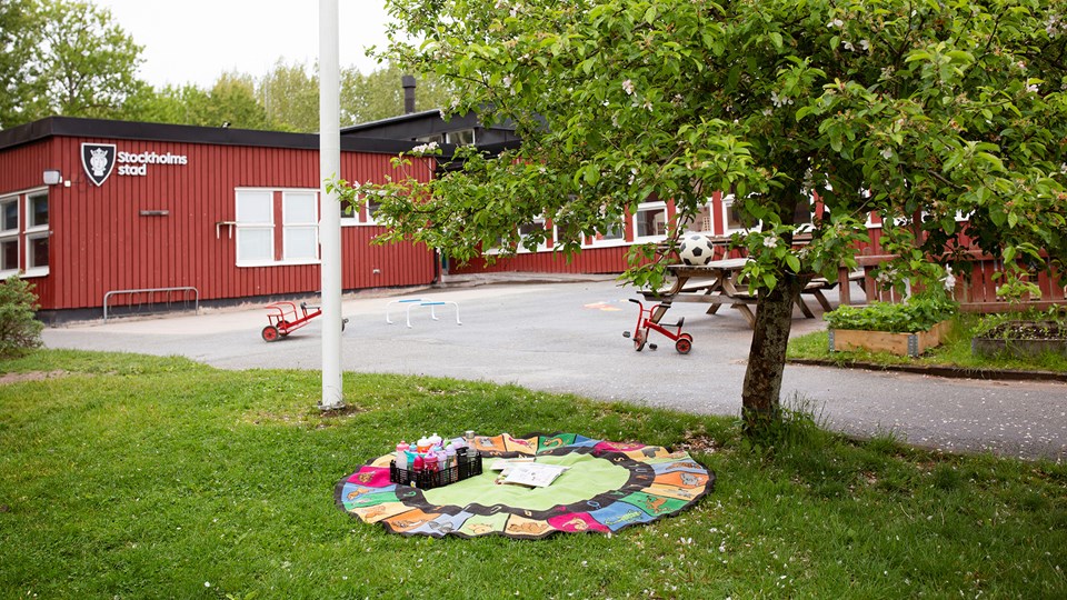 Förskolans gård med träd, planteringar, asfalt med trehjulingar och gräsmatta med leksaker.