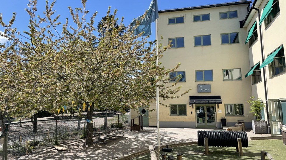 En ljusgul fyravåningsbyggnad med en gård framför som har lekverktyg och en flaggstång.