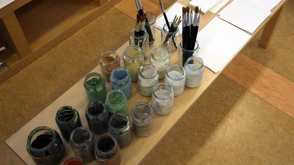 Glasburkar med färger i samt penslar på ett bord och till höger om penslarna finns vita papper.
