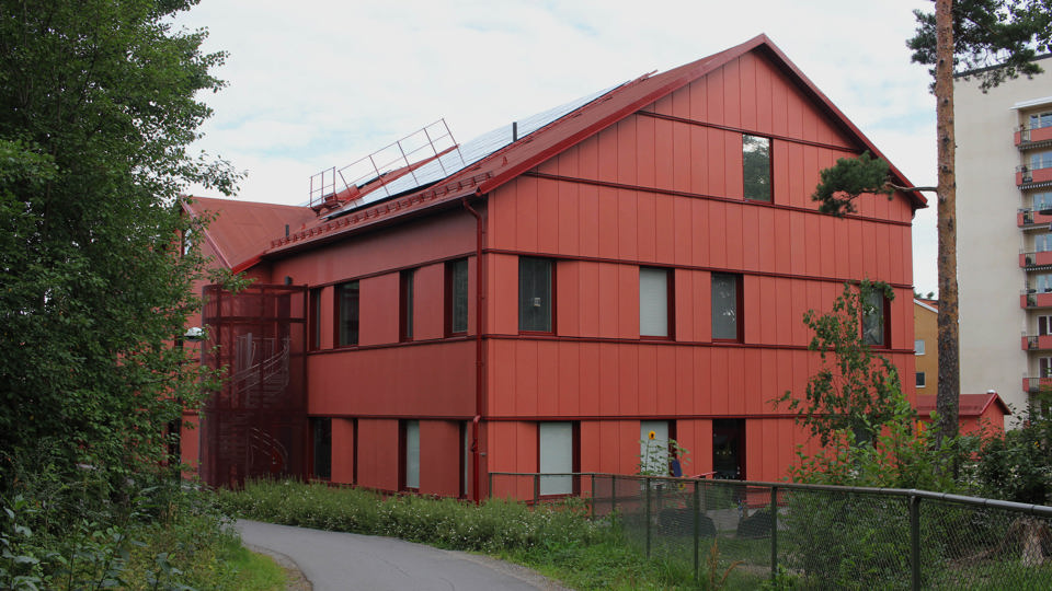 Ett rött treplanshus och en gård med buskar och träd intill en gångväg med träd intill. Bakom skymtas ett flerfamiljshus i en naturvit putsad fasad med röda balkonger.