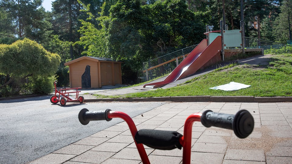 Förskolegård intill skog med asfalt, gräs och kuperad terräng på den egna gården samt rutschkana och cyklar.