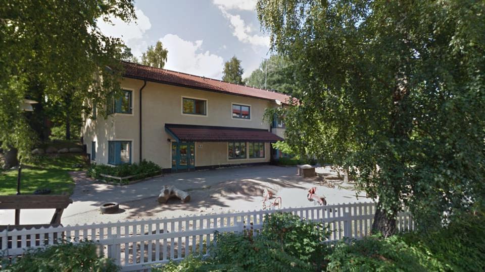 Förskolans fasad med en grå byggnad, lekplats och vita staket.